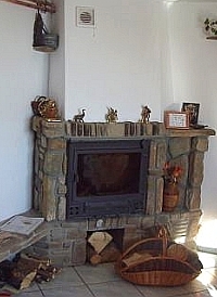 Rustykalny kominek narożny obłożony kamieniem, z dekoracjami na półce i koszem na drewno u dołu, w jasnym wnętrzu z drewnianymi akcentami.